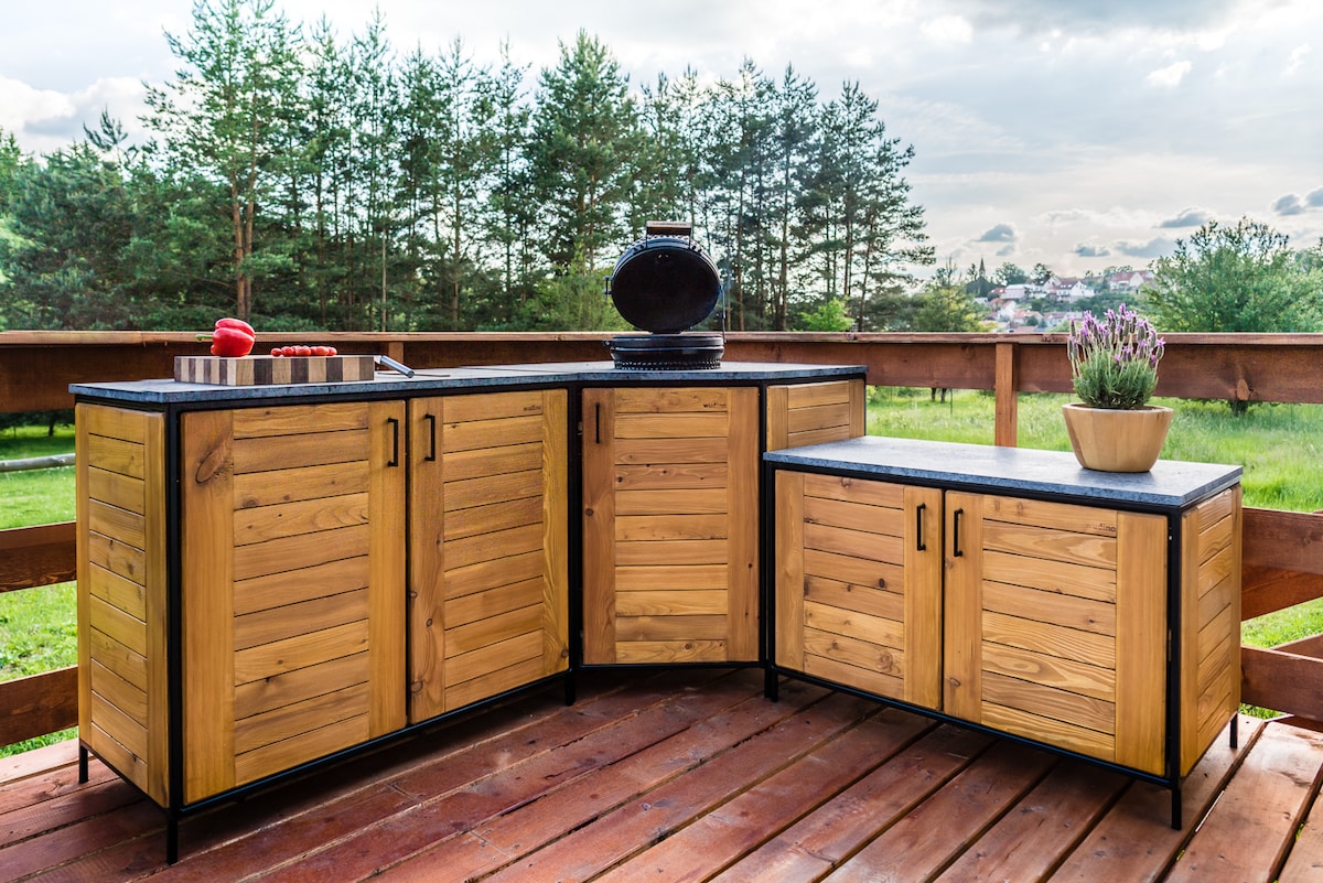 An outdoor kitchen setup features custom wooden cabinetry with a sleek countertop. The natural wood texture complements a black surface, while a decorative vase and potted plant add subtle accents. The surrounding area includes a wooden deck, offering views of lush greenery in the background.