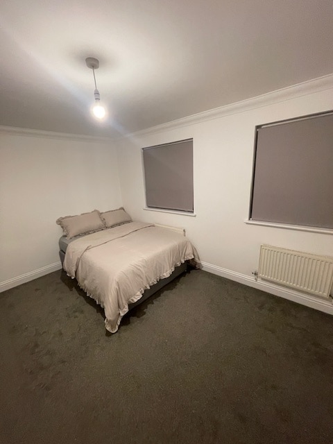 A cozy guest room is presented with a comfortable double bed adorned in soft linens. The room features two windows dressed with gray shades, allowing for natural light. A carpeted floor adds warmth to the space, complemented by a simple light fixture.