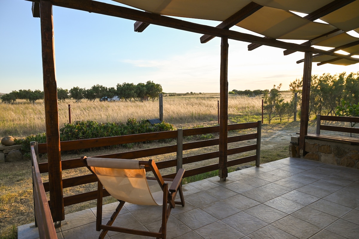 An outdoor terrace features a wooden frame with a lightweight chair positioned for viewing the surrounding landscape. The floor is tiled, and the scene is complemented by the openness of the grassy fields and a fading sunset in the distance.