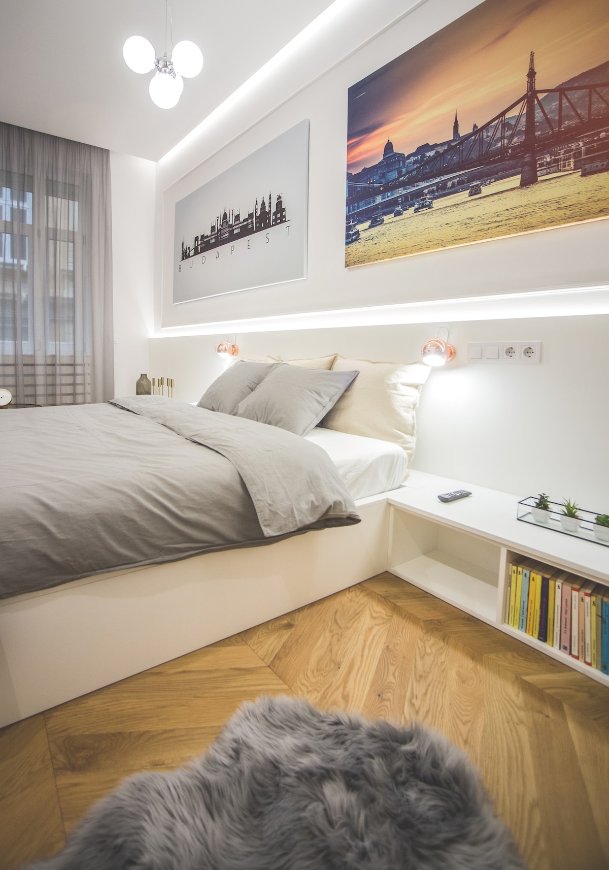 A spacious sleeping area features a large, light-colored bed with soft gray bedding. Stylish wall art depicting a cityscape hangs above. Natural light filters through sheer curtains, illuminating the wood flooring. A cozy area rug and a small nightstand with plants add to the inviting ambiance.