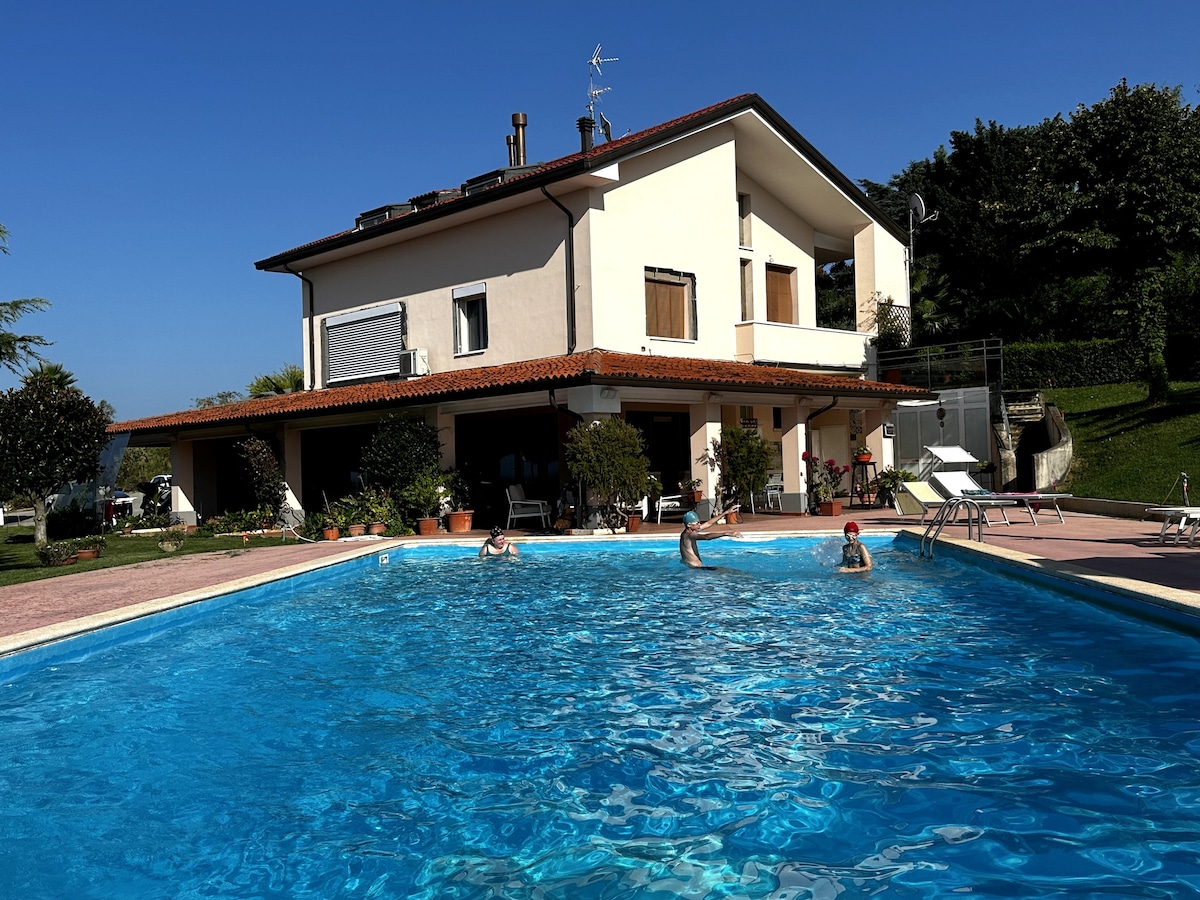 A large saltwater swimming pool is surrounded by a paved area, featuring several sun loungers and umbrellas. The villa with a terracotta roof overlooks the pool and gardens, while a clear blue sky enhances the serene atmosphere.