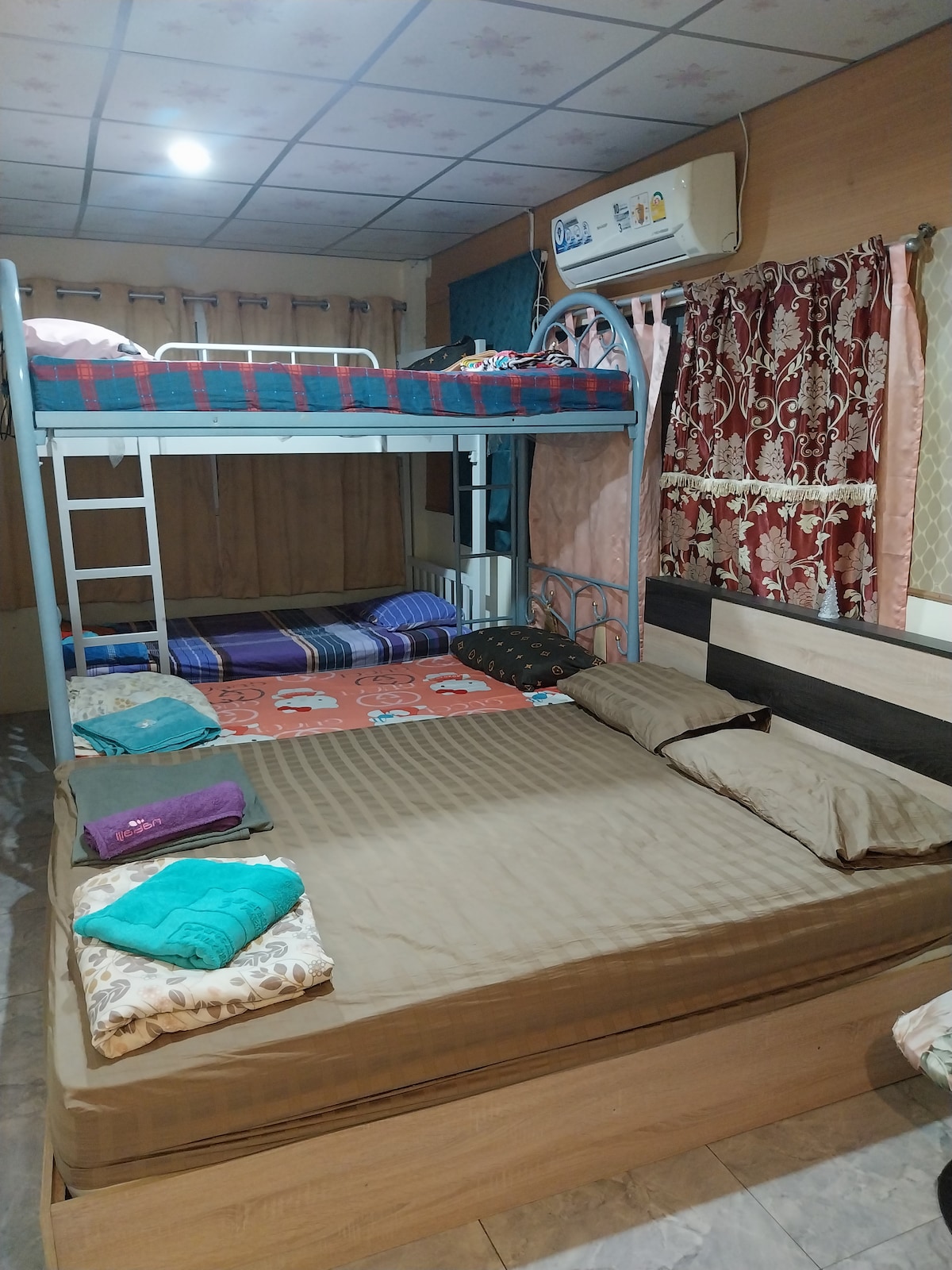 A multi-level sleeping arrangement features a bunk bed above a spacious bed. Colorful bedding in shades of blue and red is arranged neatly on each surface. Towels in various colors are placed on the larger bed, while elegant curtains frame the window, contributing to the room's overall design.