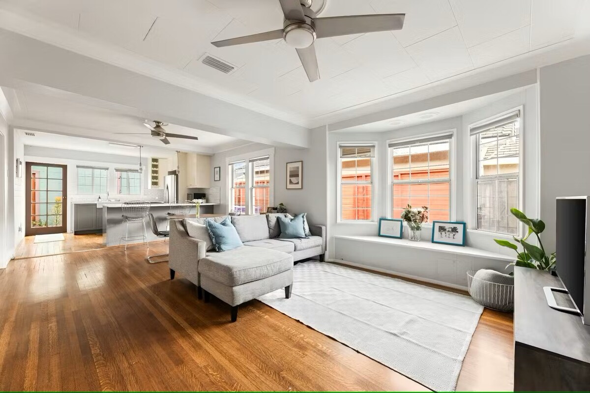 A modern living area features a light-colored sectional sofa adorned with blue cushions. Large windows allow natural light to fill the space, highlighting the hardwood floors. A white area rug lies on the floor, while a minimalist entertainment unit and potted plant add to the inviting ambiance.