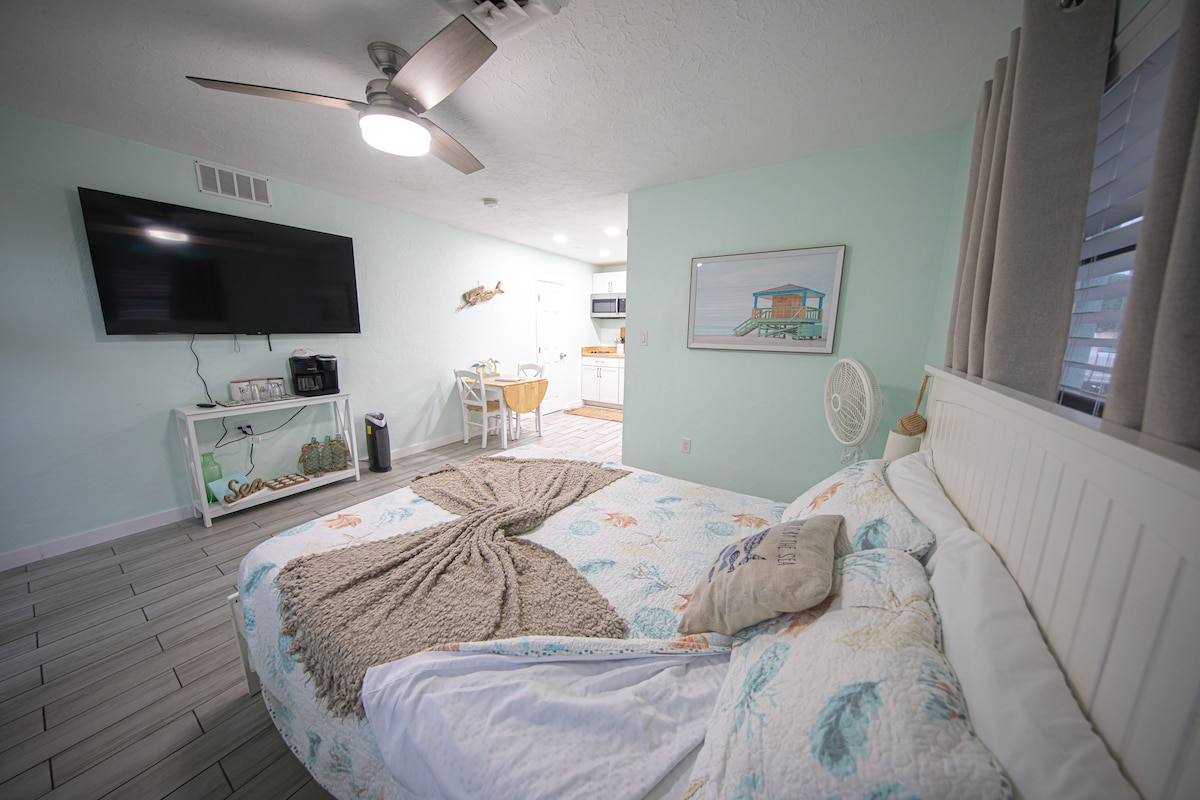 A spacious sleeping area features a queen-sized bed adorned with a patterned comforter and a cozy throw blanket. A large flat-screen TV is mounted on the wall opposite. In the background, a dining area can be seen, along with kitchen amenities.