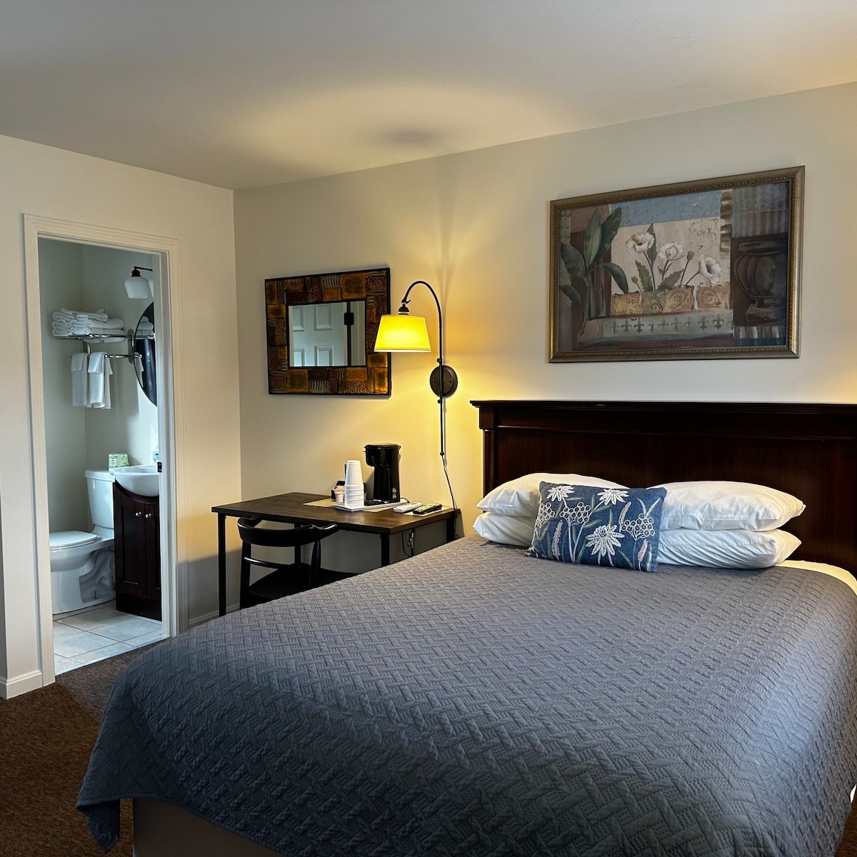 A spacious sleeping area is highlighted by a large bed with crisp bedding and a decorative blue pillow. A nightstand lamp provides soft light, while a small desk is visible nearby. A private bathroom is accessible through an interior door, featuring towels and toiletries.