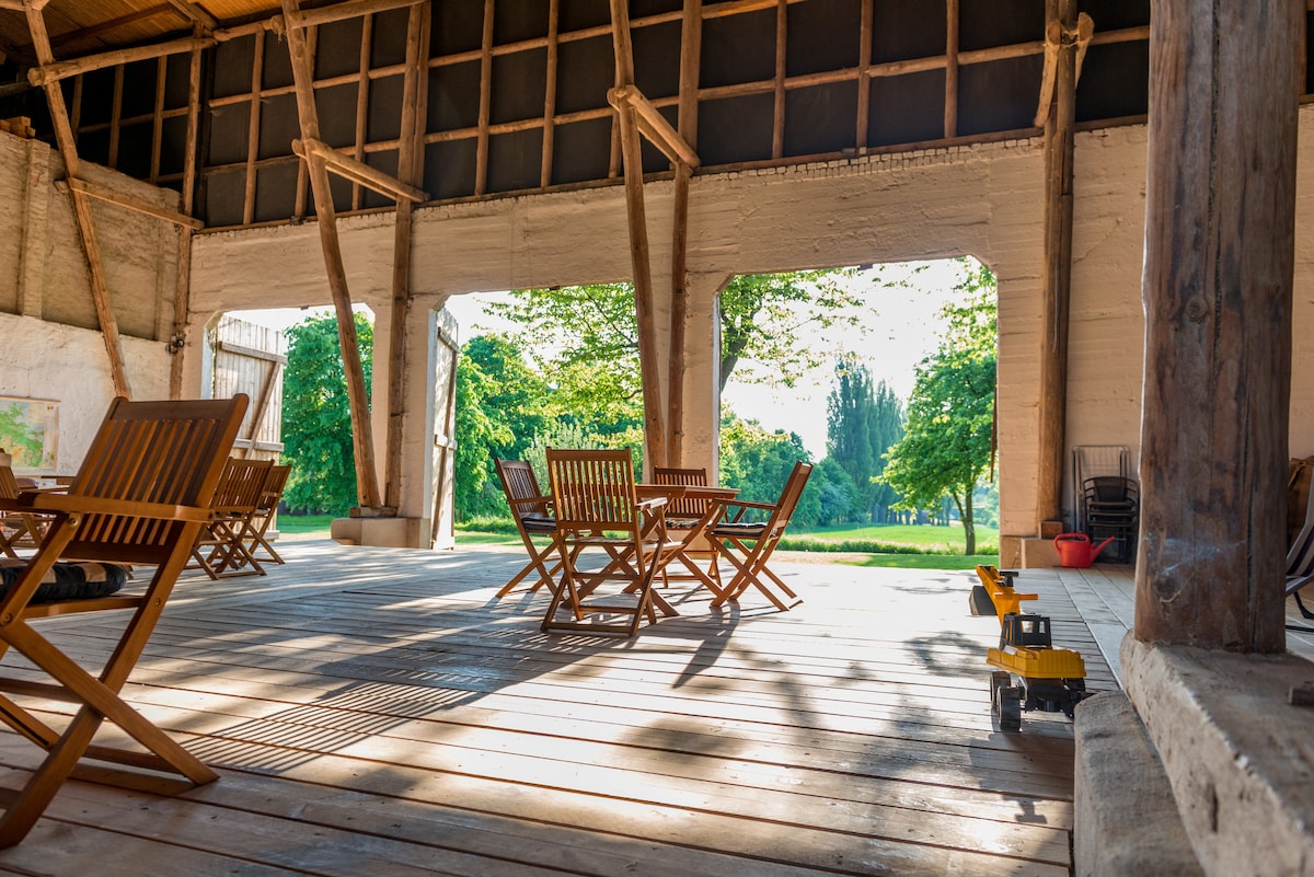 An open space is characterized by wooden beams and a smooth wooden floor, with multiple folding chairs and tables arranged for gatherings. Soft sunlight filters through large openings, illuminating the greenery outside and creating a serene environment.