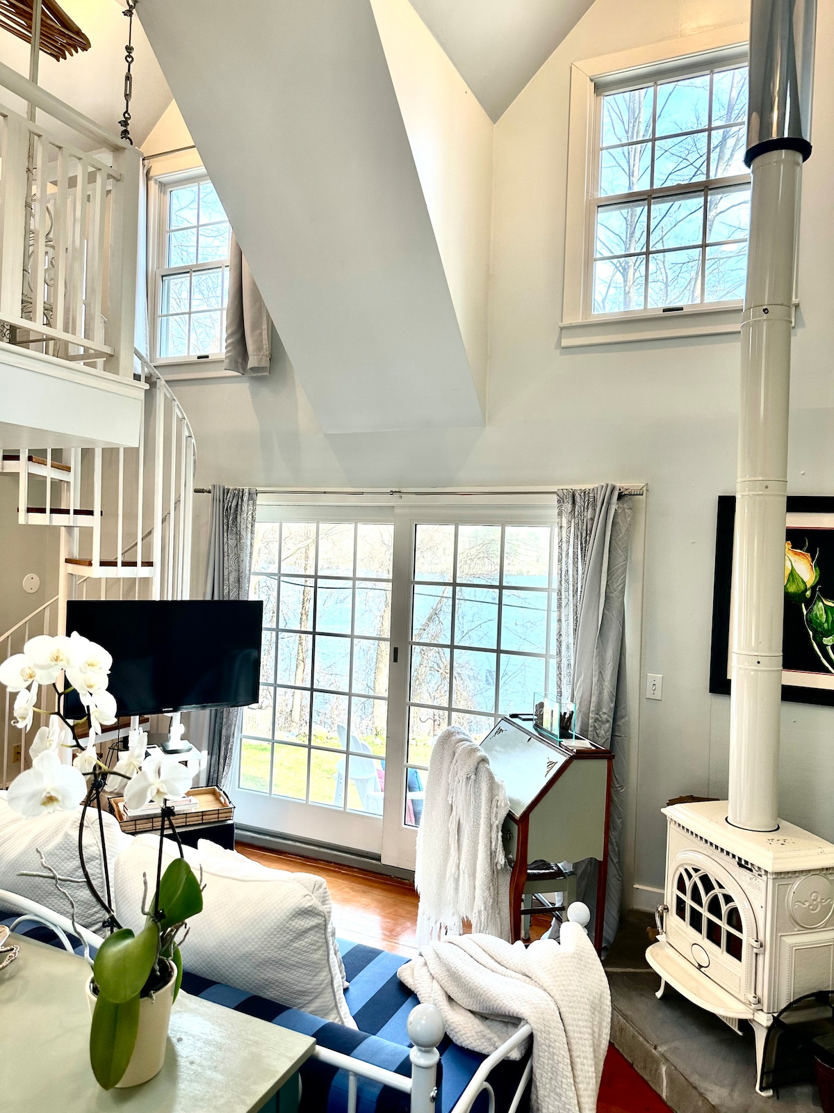 The interior features large windows allowing natural light to illuminate the space. A spiral staircase leads to the loft. A cozy seating area includes a white sofa and a decorative fireplace. Lake views are visible through the glass doors, enhancing the serene atmosphere.