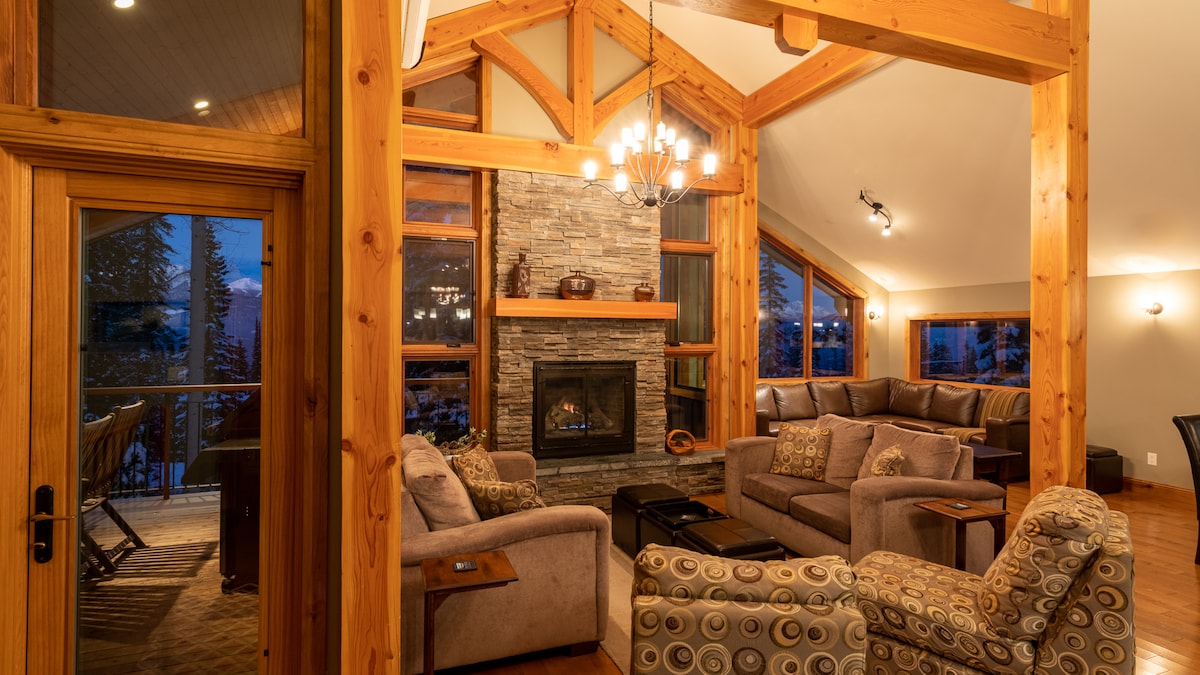 The living area features a warm color palette with comfortable seating arranged around a central stone fireplace. Large windows allow for natural light and mountain views, while the timber frame design adds an element of rustic charm. A dining area is visible through an open doorway.