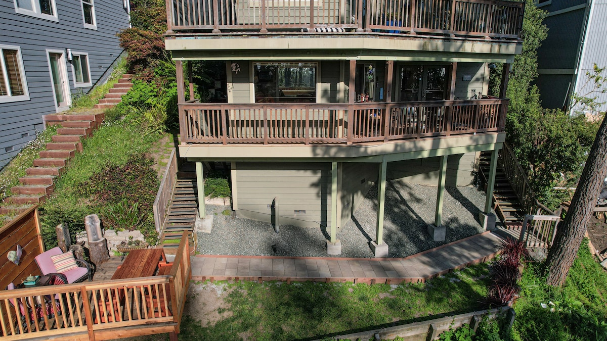The exterior of the three-story home shows a lower suite with a private entrance. A spacious patio is visible, surrounded by greenery, featuring outdoor seating. Stairs lead to the upper levels, adorned with railings, offering access to the main home.