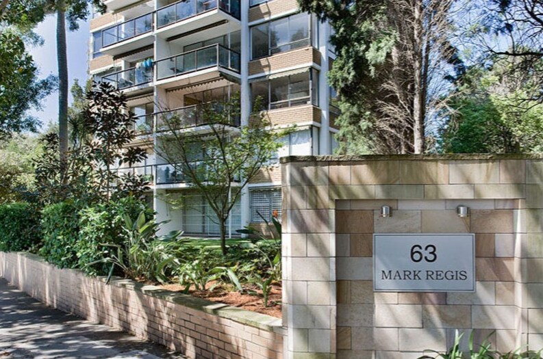 The image displays the front of the seven-storey security building, featuring a modern facade with balconies overlooking the greenery. A well-maintained garden is visible, offering a blend of plants and trees. The property address is clearly indicated on a stone wall.