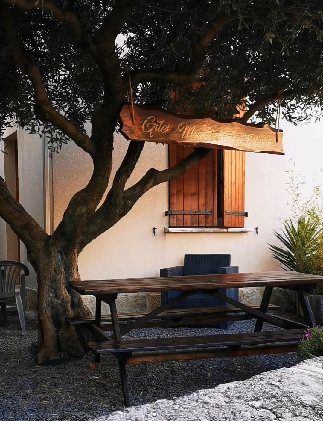 An outdoor setting is highlighted, featuring a large wooden table under a tree. A sign hangs above, displaying the name of the gîte. A chair is positioned near the table, and a window is visible in the background, contributing to a tranquil outdoor space.