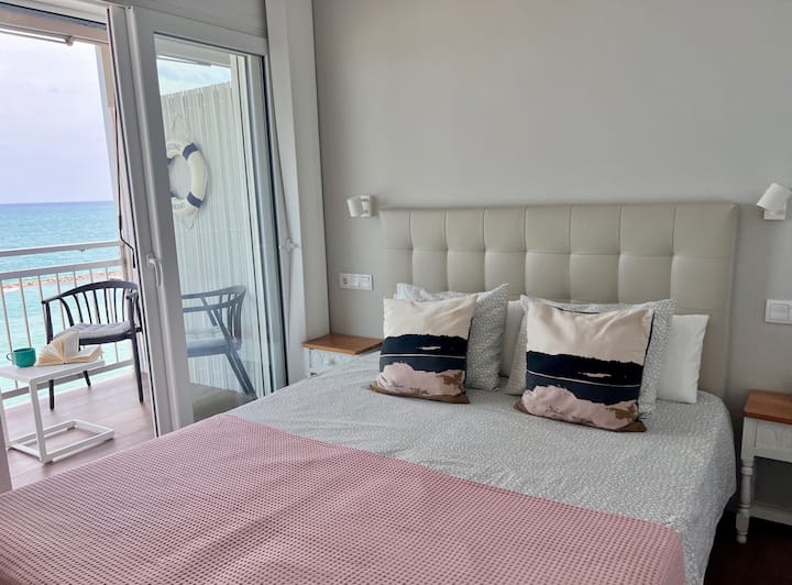 Dormitorio principal con acceso directo a la terraza. La cama queen size, flanqueada por suaves luces de noche con las que poder relajarse leyendo, invita al descanso mientras el mar se asoma por el ventanal creando un ambiente sereno y mediterráneo.