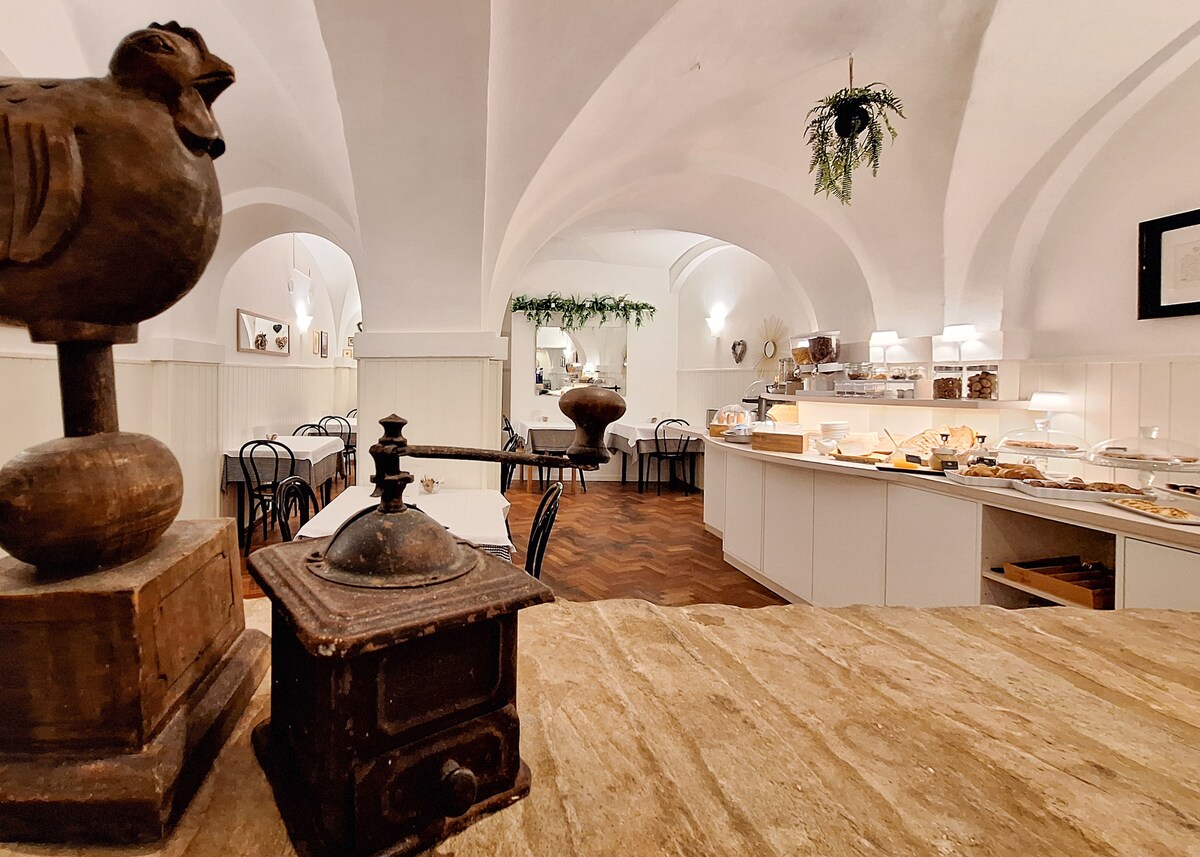 An inviting breakfast area features traditional wooden decor and a variety of baked goods displayed on a long counter. Arched ceilings create a spacious atmosphere, complemented by soft lighting and elegant furnishings. Decorative elements, including a small urn and a vintage grinder, add character to the setting.