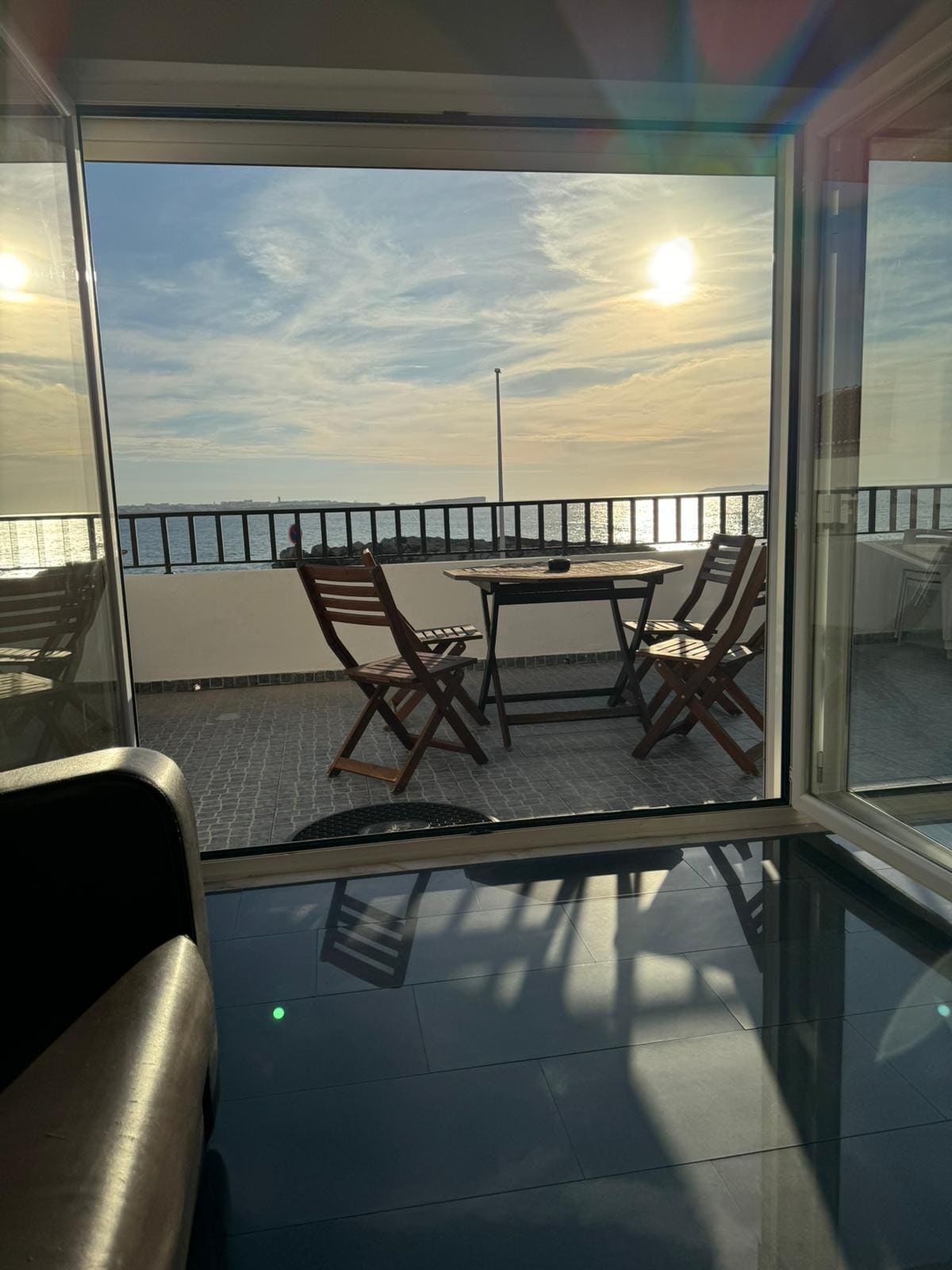 A sunlit terrace features two wooden chairs and a small table, facing expansive views of the water. Large glass doors slide open, connecting the indoor space with the outdoor area. The floor reflects the sunlight, enhancing the open atmosphere.