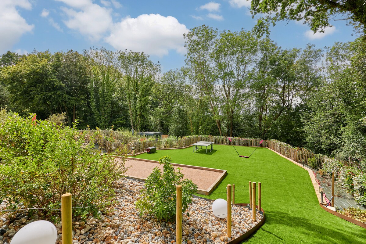A shared garden area is shown, featuring a well-maintained lawn and gravel paths. Various plants and trees create a natural backdrop, while a petanque court and lawn for games are visible. Soft lighting elements are placed throughout the space, enhancing the garden's tranquil ambiance.