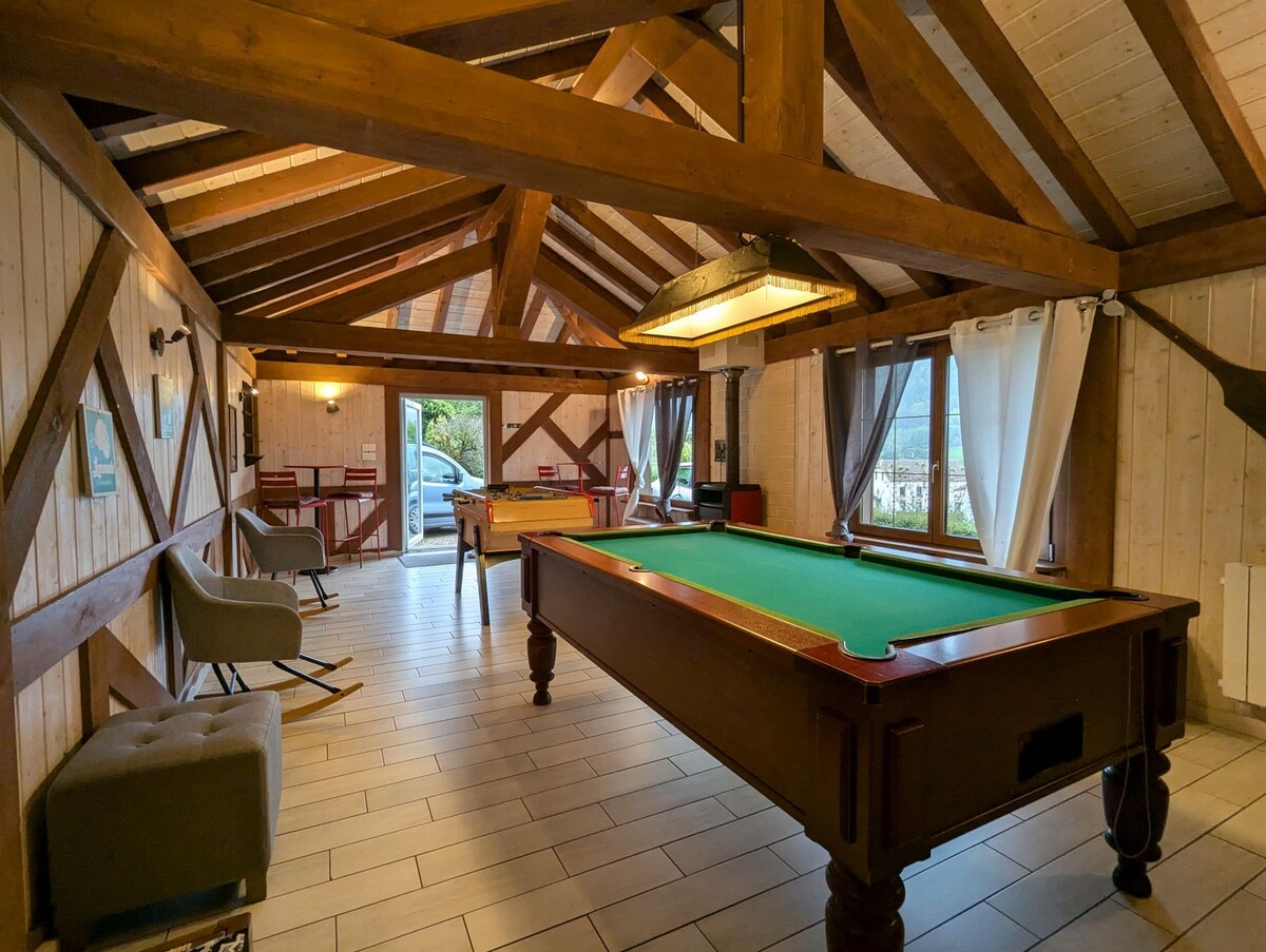 A spacious game room features a billiard table, surrounded by wooden beams and natural light filtering through large windows. Comfortable seating areas are arranged nearby, with decor highlighting the rustic charm. A glimpse of the outdoors is visible through partially drawn curtains.