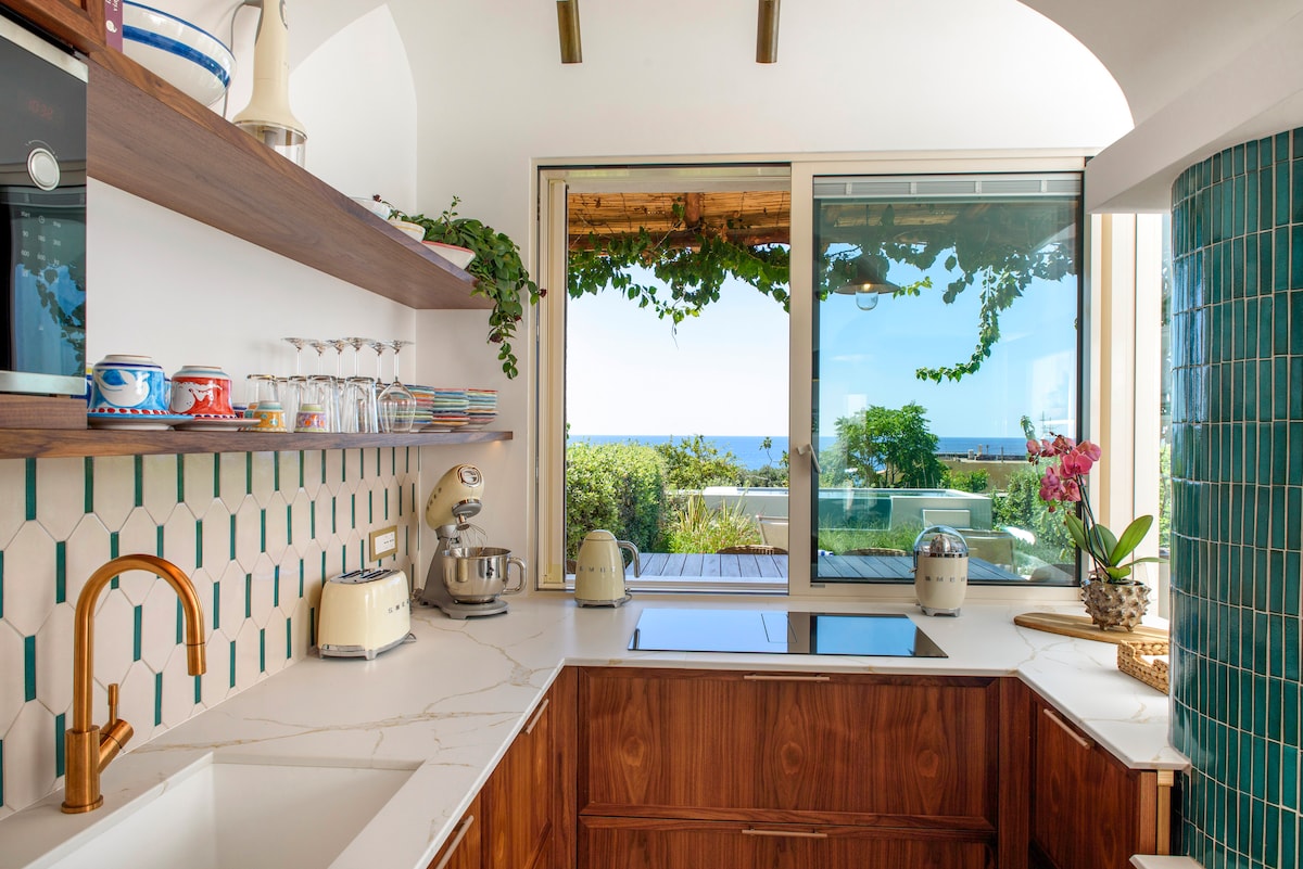 A modern kitchen space features a marble countertop and wooden cabinetry. Glassware and cooking essentials are neatly arranged. A large window allows natural light to fill the room, providing a view of the garden and the sea in the distance.