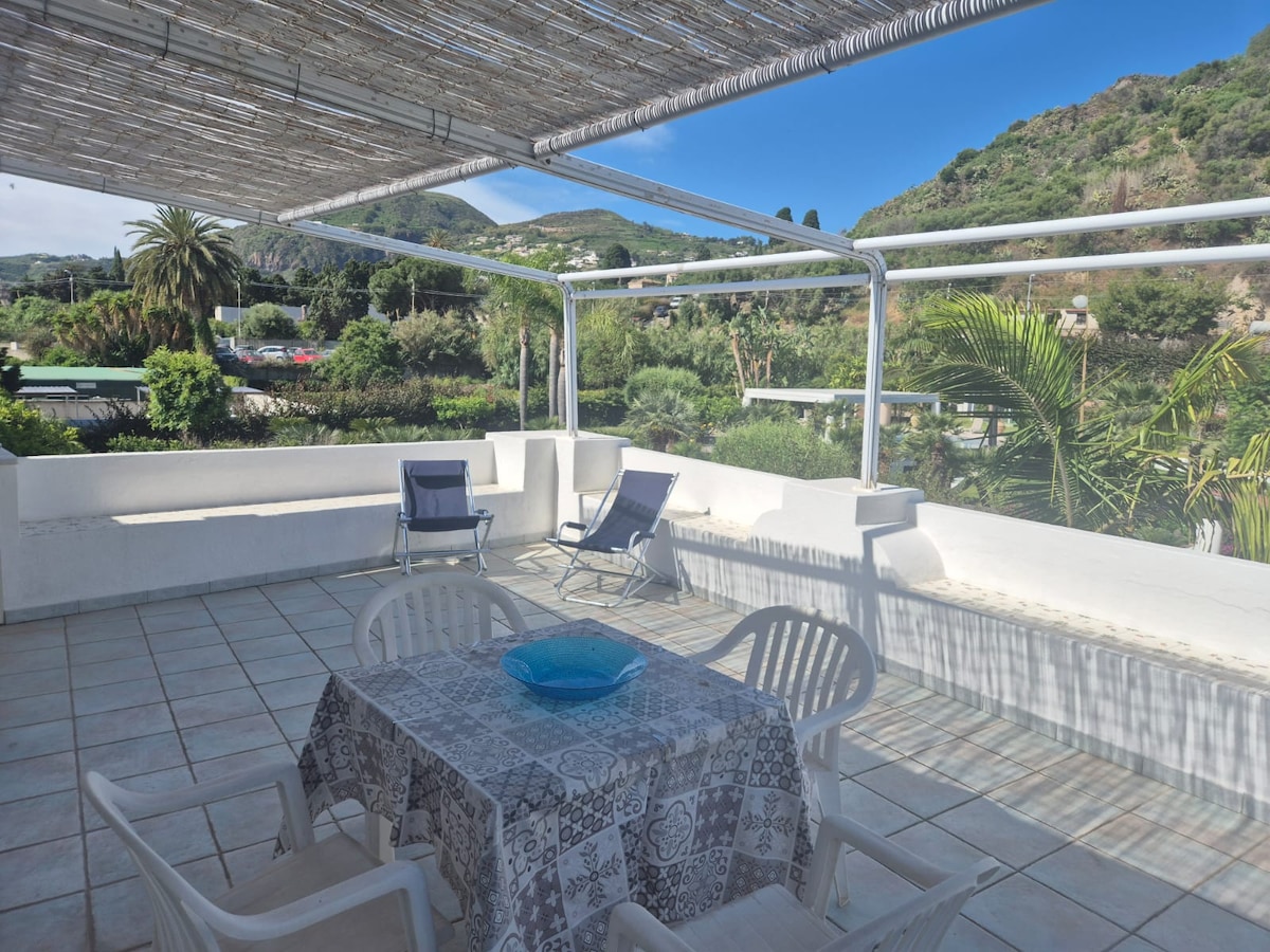 A spacious terrace is highlighted by a light-colored tiled floor. A rectangular table is set for outdoor dining, surrounded by four white chairs. Lounge chairs are positioned for relaxation, with lush greenery visible in the background and mountains creating a serene backdrop.