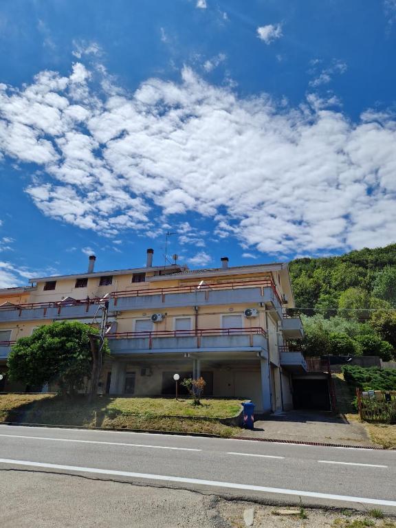 A three-story building is seen under a bright blue sky with scattered clouds. Green hills rise in the background, providing a natural backdrop. Large balconies with railings are visible on the upper floors, along with individual air conditioning units mounted on the exterior.