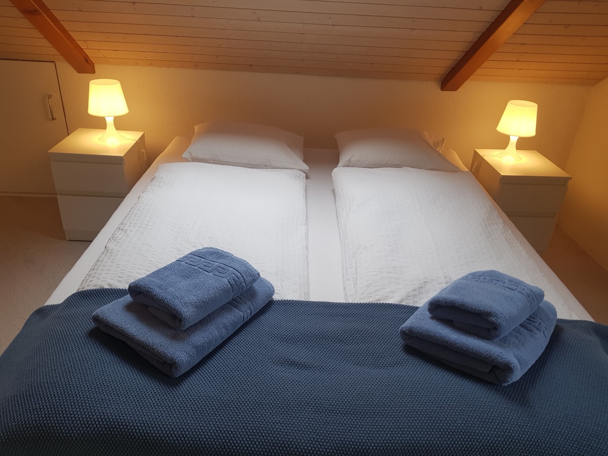 A cozy bedroom is presented with two spacious beds, each adorned with white linens. Two bedside lamps provide soft lighting, and neatly folded towels are arranged on a dark blue blanket at the foot of the bed. Wooden beams add warmth to the ceiling.