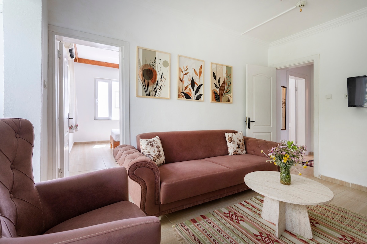 A relaxed living area features a plush brown sofa with decorative pillows and a round coffee table. Artful wall decorations with natural themes enhance the space. Light-colored walls are complemented by a soft area rug, and a doorway leads to additional rooms, inviting exploration.