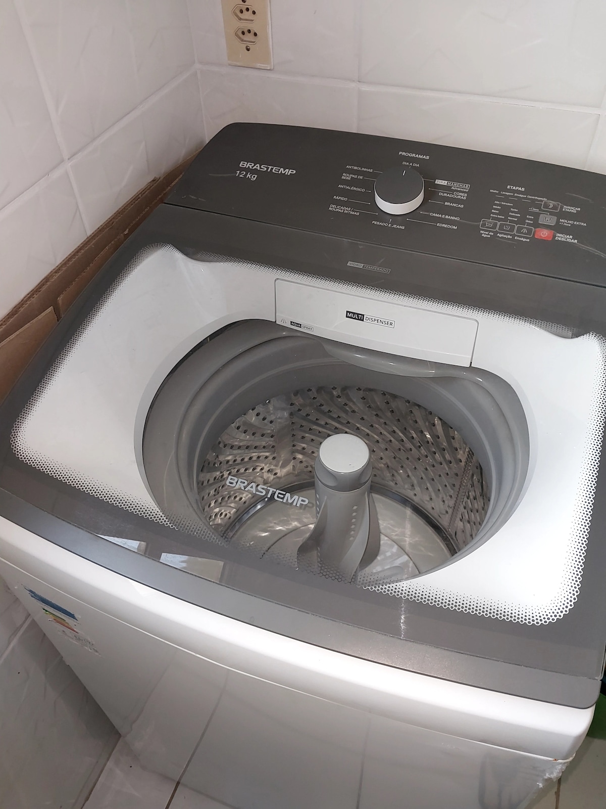 A modern washing machine is displayed, featuring a transparent top lid and a visible control panel with various settings. The interior drum is stainless steel, and the exterior is finished in white with gray accents, set against tiled walls.