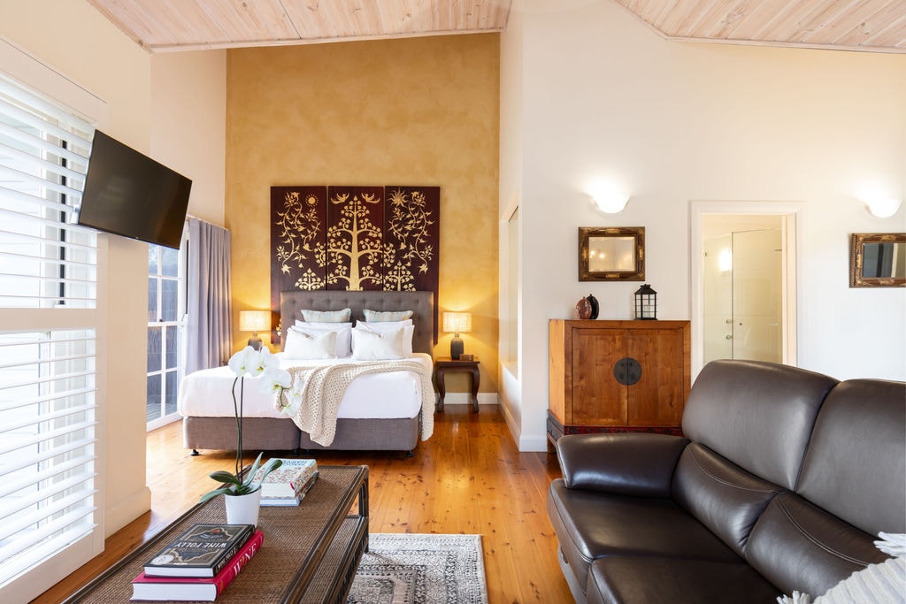 An open-plan villa showcases a spacious bedroom area with a king-size bed, complemented by textured wall art and warm lighting. Nearby, a comfortable leather lounge faces a decorative coffee table, while large windows provide natural light and botanical views. Soft tones enhance the inviting atmosphere.