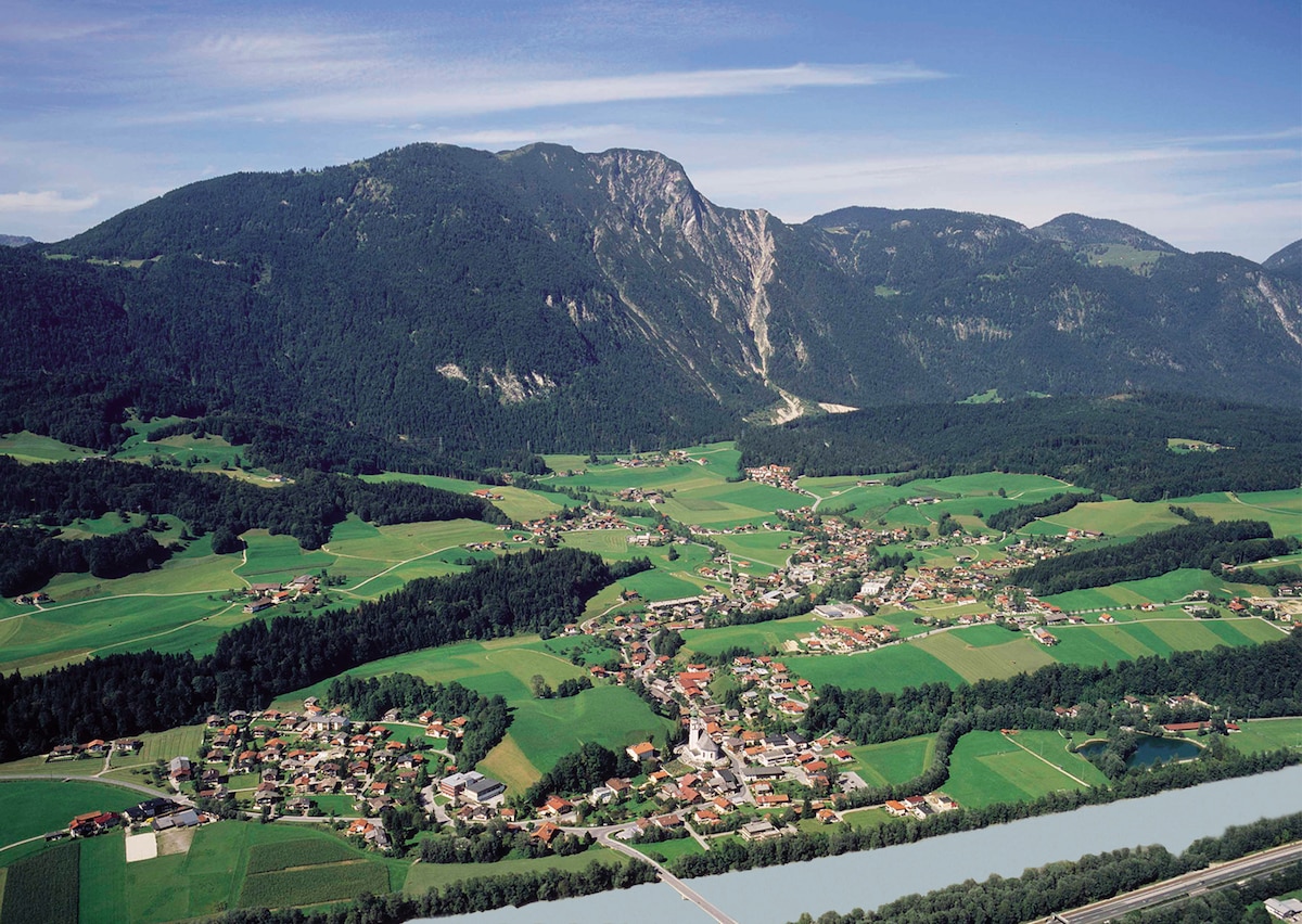 Aerial view of a rural landscape featuring a small village nestled among rolling hills and lush green fields. A river runs along the bottom of the image, complementing the vibrant scenery. Mountains rise in the background, contributing to the serene natural environment.