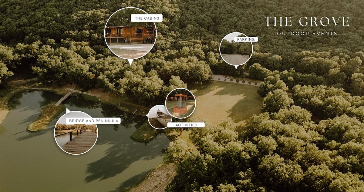 An aerial view showcases a lush, wooded area surrounding a tranquil pond. Wooden structures for cabins are visible, along with designated parking spaces. Pathways lead to outdoor activity areas, including a bridge that connects to a small peninsula, offering a natural and serene setting.