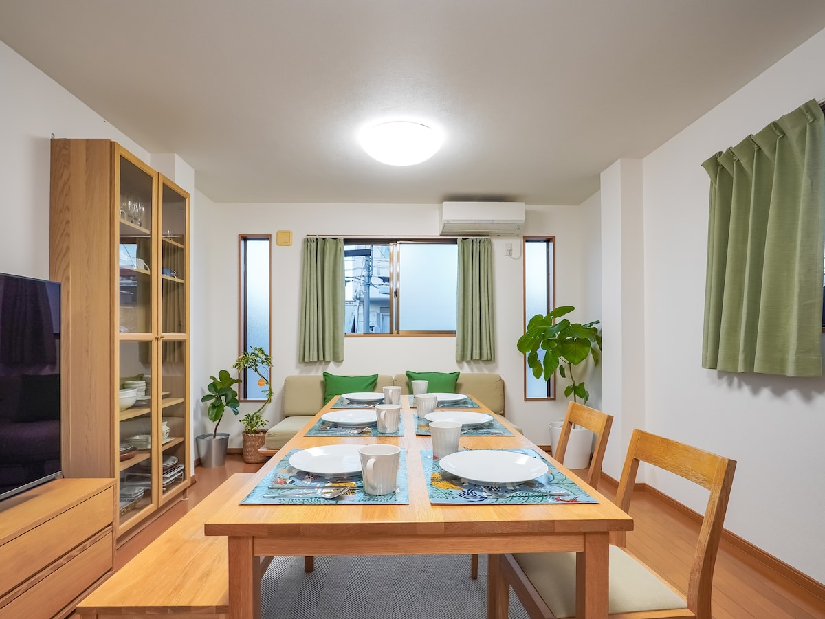 A dining area is presented with a wooden table, set for six with plates, cups, and napkins. Green cushions are arranged on the chairs, and large windows allow natural light to fill the space. A tall plant adds a touch of greenery to the room.