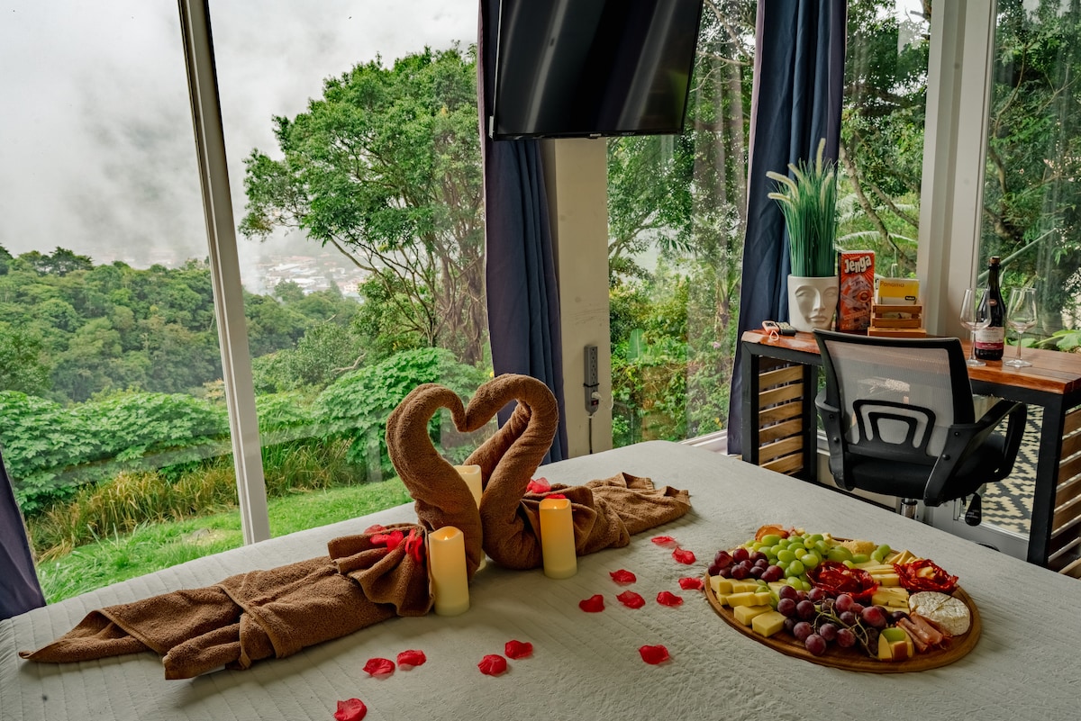 A heart-shaped arrangement of towels is placed on the bed, accompanied by a platter of fresh fruits. Candles and rose petals enhance the scene. Large windows showcase expansive views of the lush greenery outside, while a desk with a chair is positioned nearby.