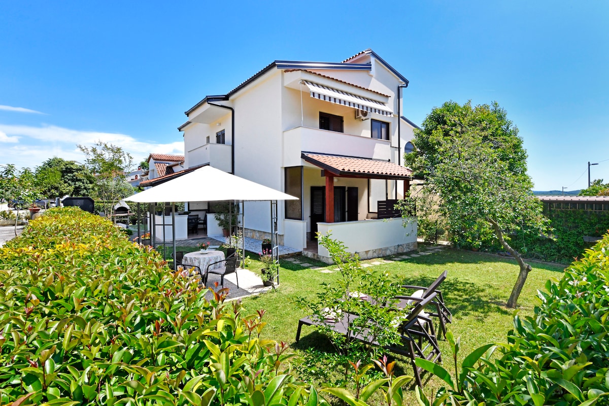 The exterior of the family house features a well-maintained garden bordered by lush greenery. A shaded seating area is visible under a canopy, with a table and chairs arranged for relaxation. A glimpse of the home’s upper balcony can also be seen.