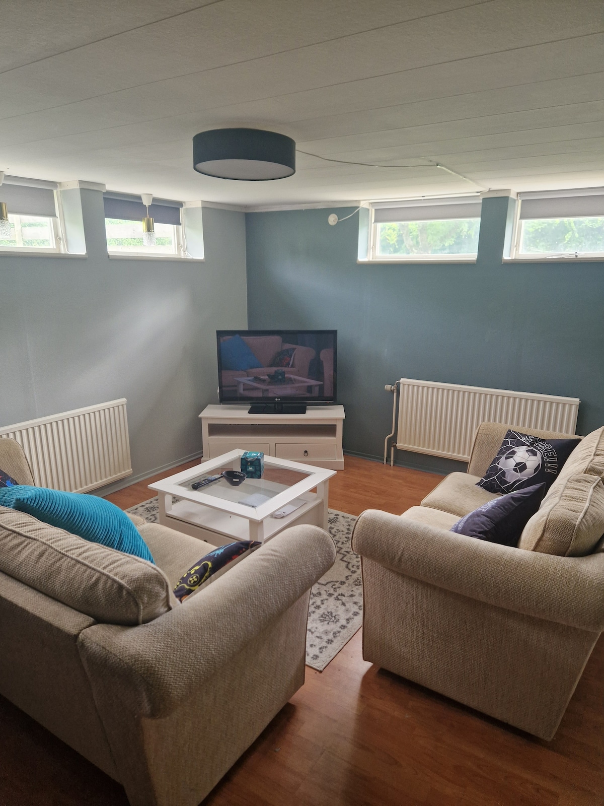 A cozy seating area is furnished with two light-colored sofas and a white coffee table. A television is positioned against a wall, accompanied by decorative items. Natural light enters through multiple windows located near the ceiling, enhancing the room's atmosphere.