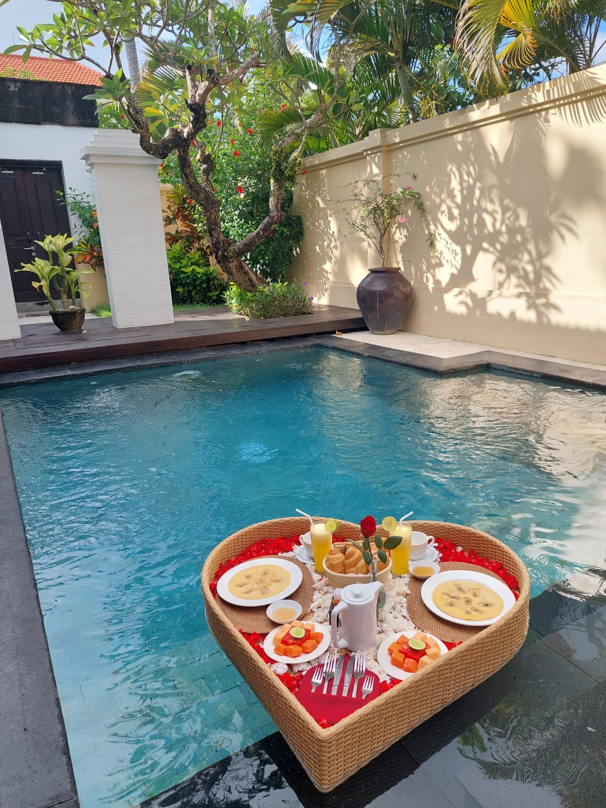 A heart-shaped basket is positioned on the water's surface of a private pool, featuring an assortment of breakfast items, beverages, and garnishes. Lush greenery and tropical plants are visible in the background, complemented by a decorative vase beside the pool.