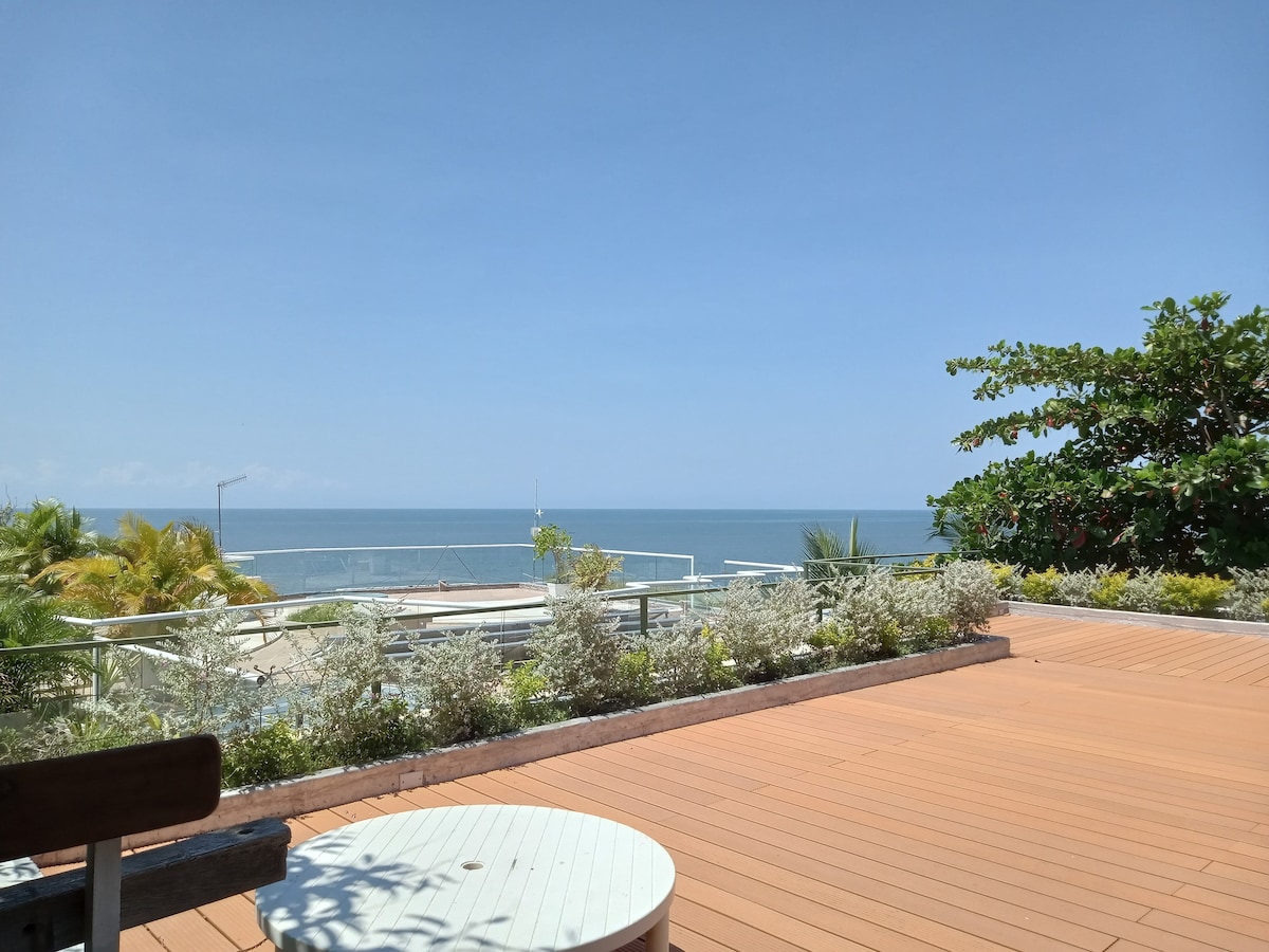An expansive outdoor terrace showcases a serene ocean view under a clear blue sky. Lush greenery and well-maintained shrubs border the space, contributing to a refreshing atmosphere. A circular table positioned on the wooden decking invites moments of relaxation.