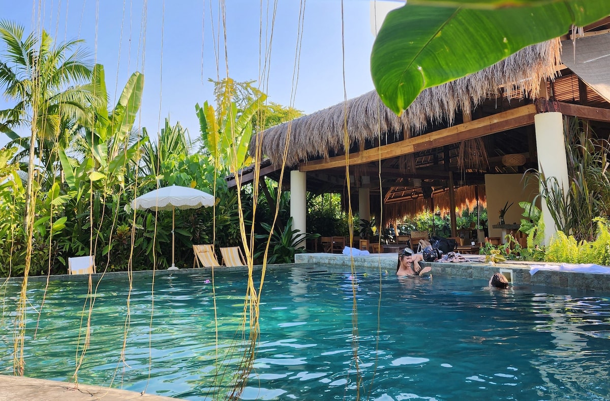 A serene pool area is surrounded by lush greenery, with several palm trees and tropical plants contributing to a natural ambiance. The inviting water reflects sunlight under a clear blue sky. A thatched-roof structure provides shaded seating options nearby.