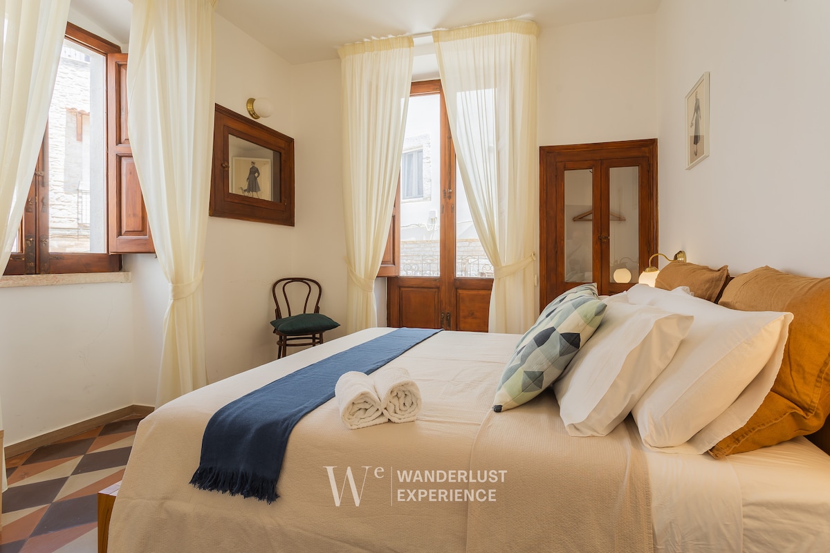 A bright bedroom features soft natural light filtering through layered curtains. A spacious bed is adorned with neutral-toned linens, complemented by decorative pillows. A chair sits in the corner next to a window, with a small table visible in the background. Wooden framed artworks add character.