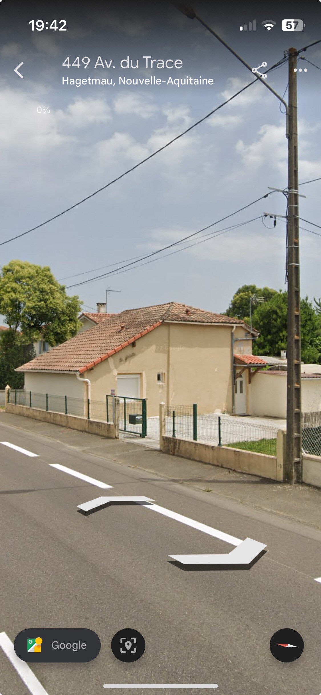 The image shows a single-story house with a light-colored exterior and a tiled roof. A paved driveway is visible in front of the house, bordered by a low fence. Trees provide greenery in the vicinity, complementing the suburban setting.