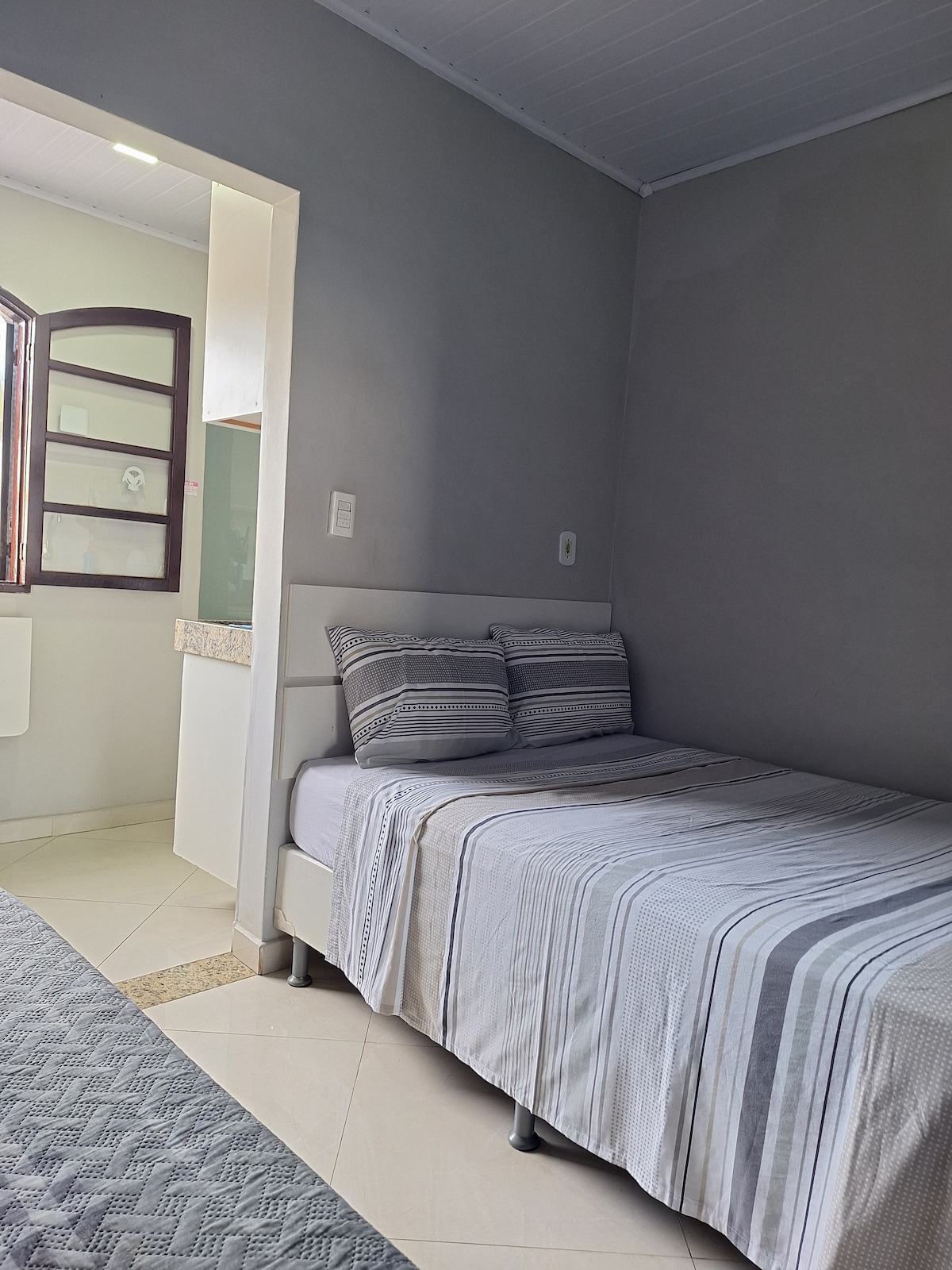 A simple sleeping area features a bed with neutral-toned striped bedding, complemented by two pillows. A window allows natural light to illuminate the space, and a glimpse of a kitchen area is visible through an open doorway.