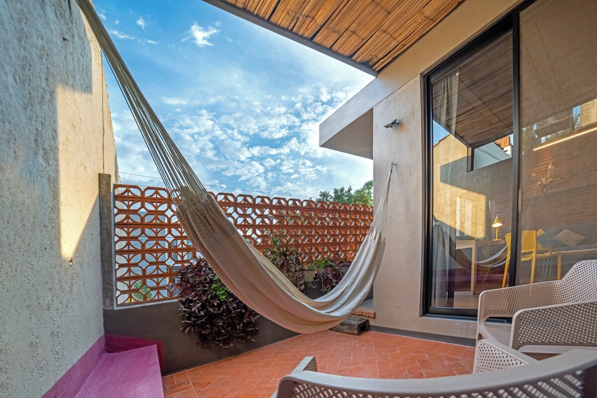 A shaded terrace features a comfortable hammock suspended between two walls, inviting relaxation. Soft natural light filters through decorative latticework, illuminating the surrounding plants. Nearby, modern seating arrangements consist of light-colored chairs, enhancing the tranquil outdoor space.