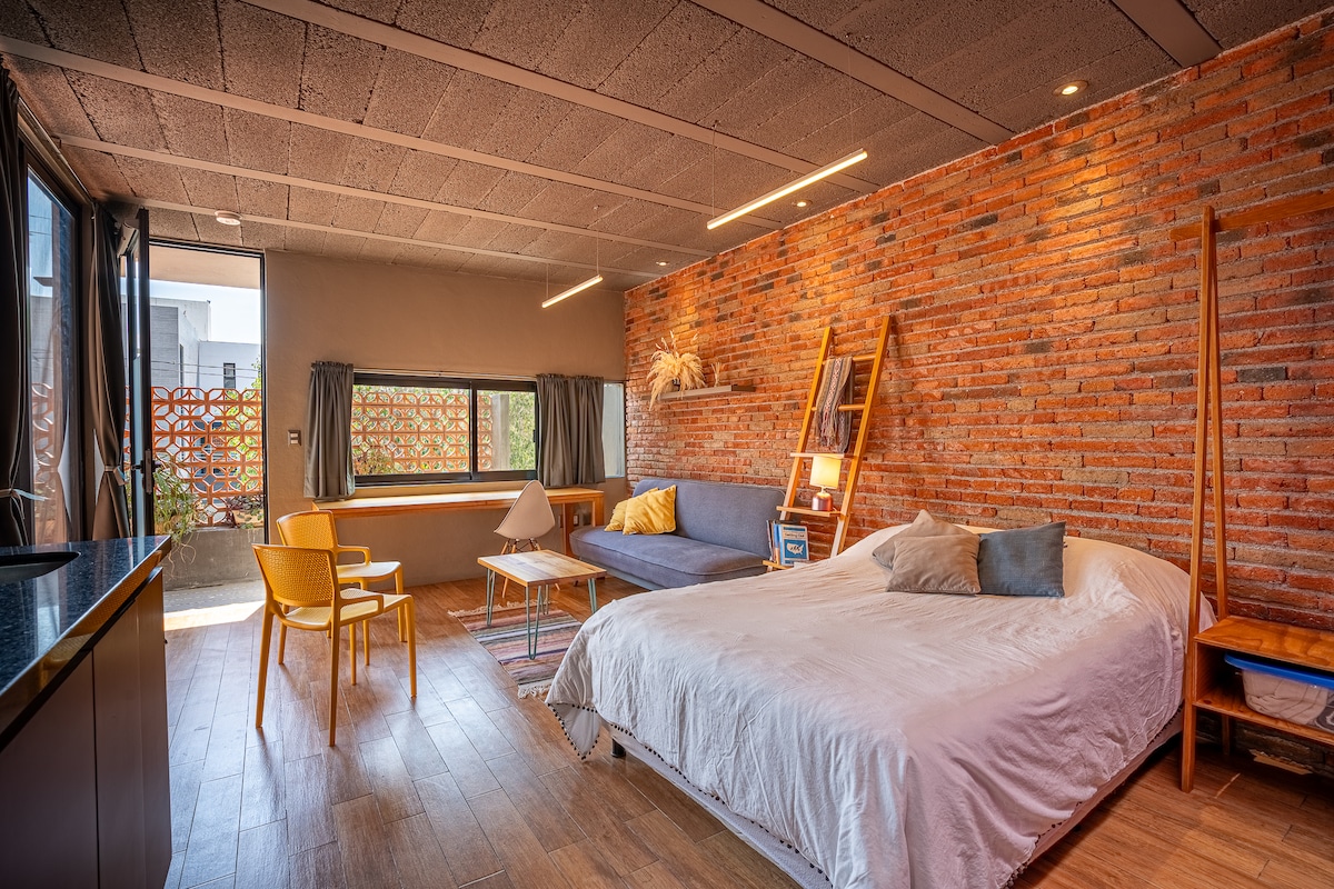 A cozy loft space features a spacious bed with light bedding, complemented by a sofa and wooden chairs. Exposed brick walls add texture, while large windows provide natural light. A simple table is positioned between the seating area, enhancing the comfort of the environment.