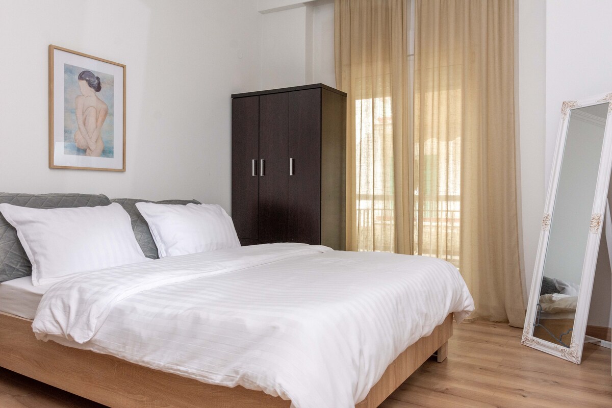 A spacious sleeping area is presented with a light-toned wooden bed dressed in white linens. A framed artwork adorns the wall, while a tall wardrobe stands beside a large window covered with soft, sheer curtains. A full-length mirror complements the decor.