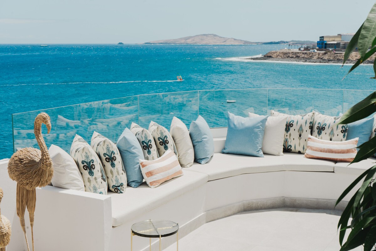 A spacious outdoor seating area is presented with a soft, curved couch adorned with various patterned cushions in light colors. The inviting view of the ocean is visible in the background, while decorative elements, including a tall decorative figure, enhance the tranquil space.
