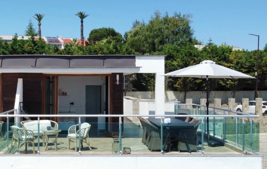 An outdoor terrace features a glass railing, offering a clear view of the surrounding greenery. A dining table and chairs are arranged under a large umbrella, providing shade. The area is well-lit by natural sunlight, creating a welcoming space for relaxation.