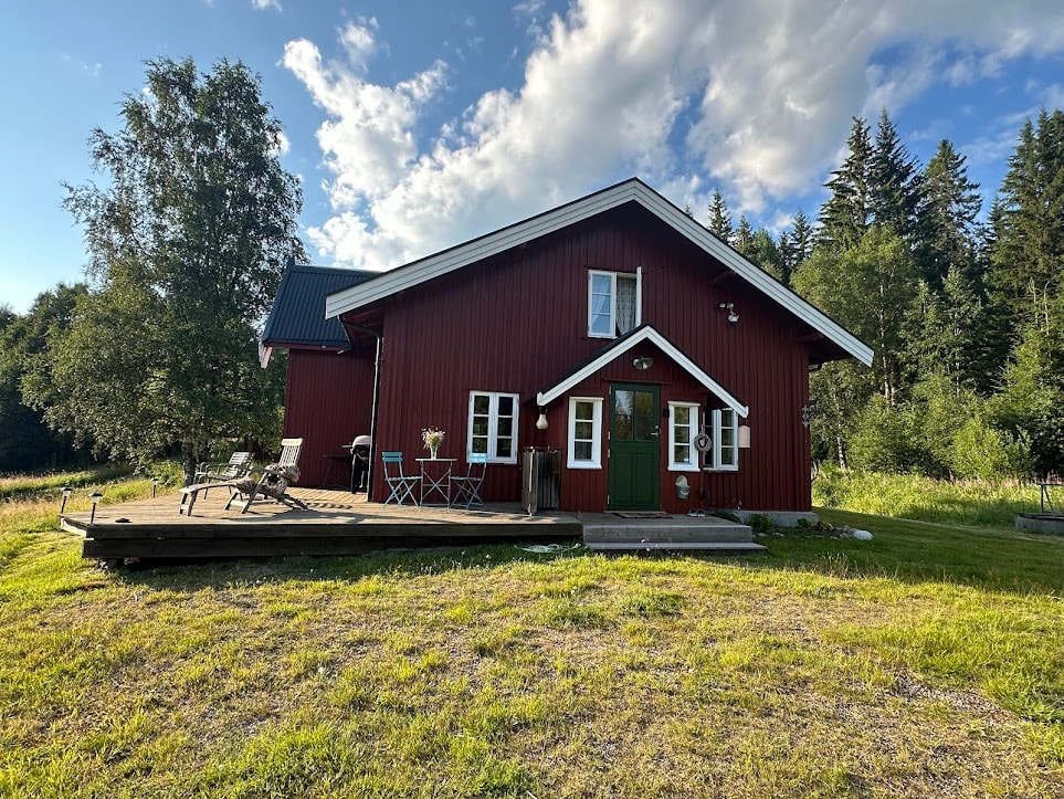 A red wooden cabin is set in a grassy area, surrounded by trees. A spacious deck extends from the cabin, furnished with chairs and a small table. Large windows allow natural light to illuminate the interior, which is complemented by a welcoming green door.