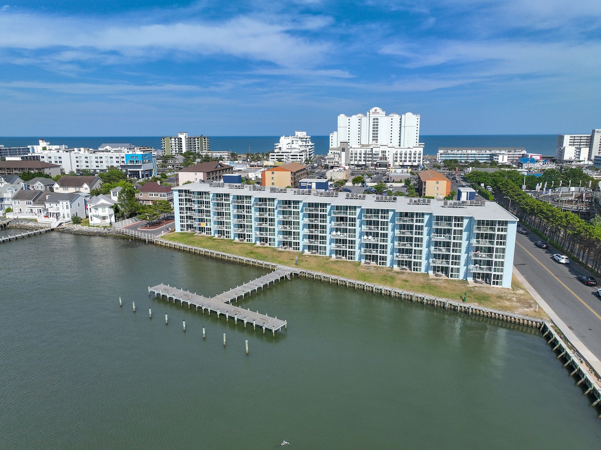 Aerial view of a bayfront condo building is presented, showcasing multiple balconies overlooking the water. The surrounding area includes a tranquil bay and nearby establishments, with the ocean visible in the distance under a clear blue sky.
