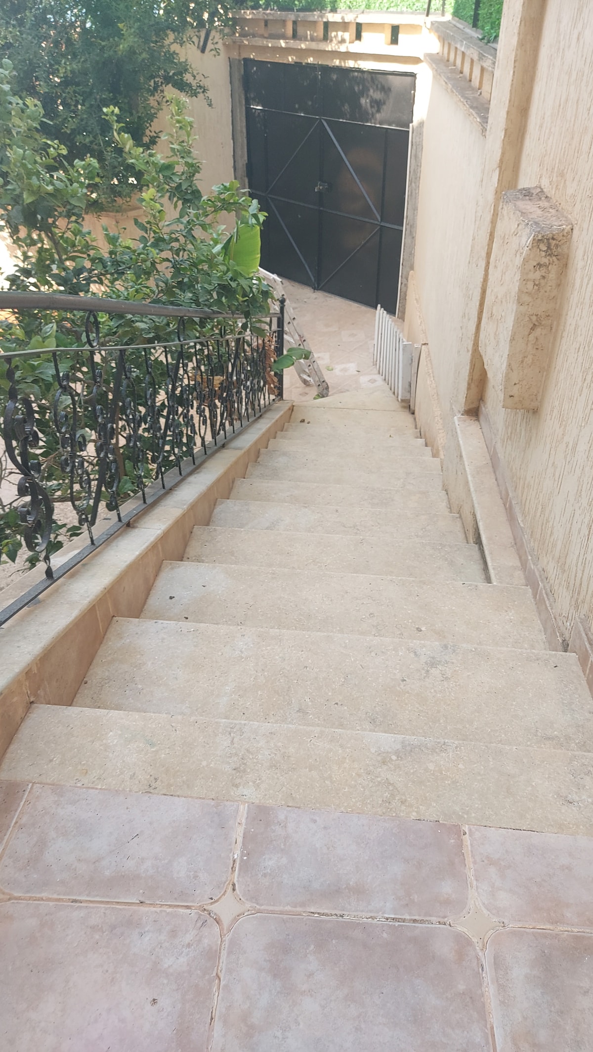 A stone staircase is seen descending towards a gated area. The surrounding walls are textured and light beige, with a decorative iron railing visible along one side. Green foliage is observed on the left, contributing to the serene residential ambiance.
