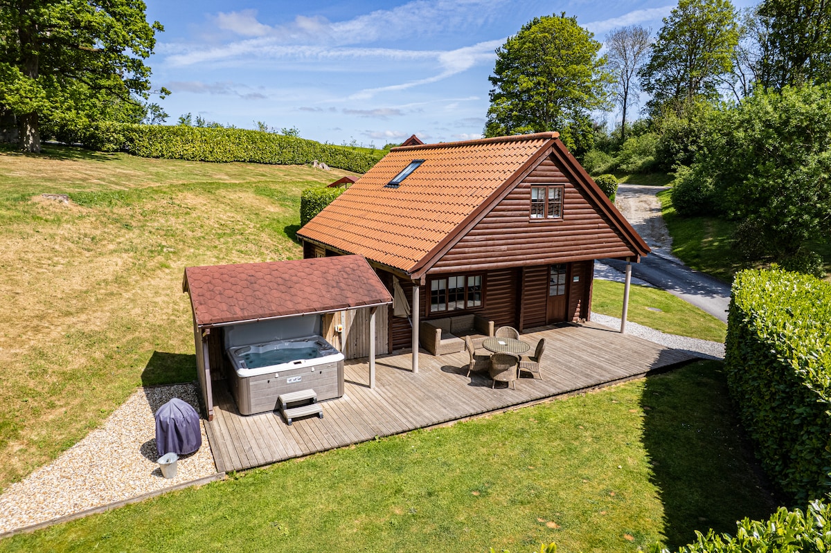 A wooden lodge is set within a grassy area, featuring a deck with outdoor seating and a hot tub. Lush green trees and a gravel pathway frame the setting. The red-tiled roof and windows allow for natural light, creating a harmonious blend with the surroundings.