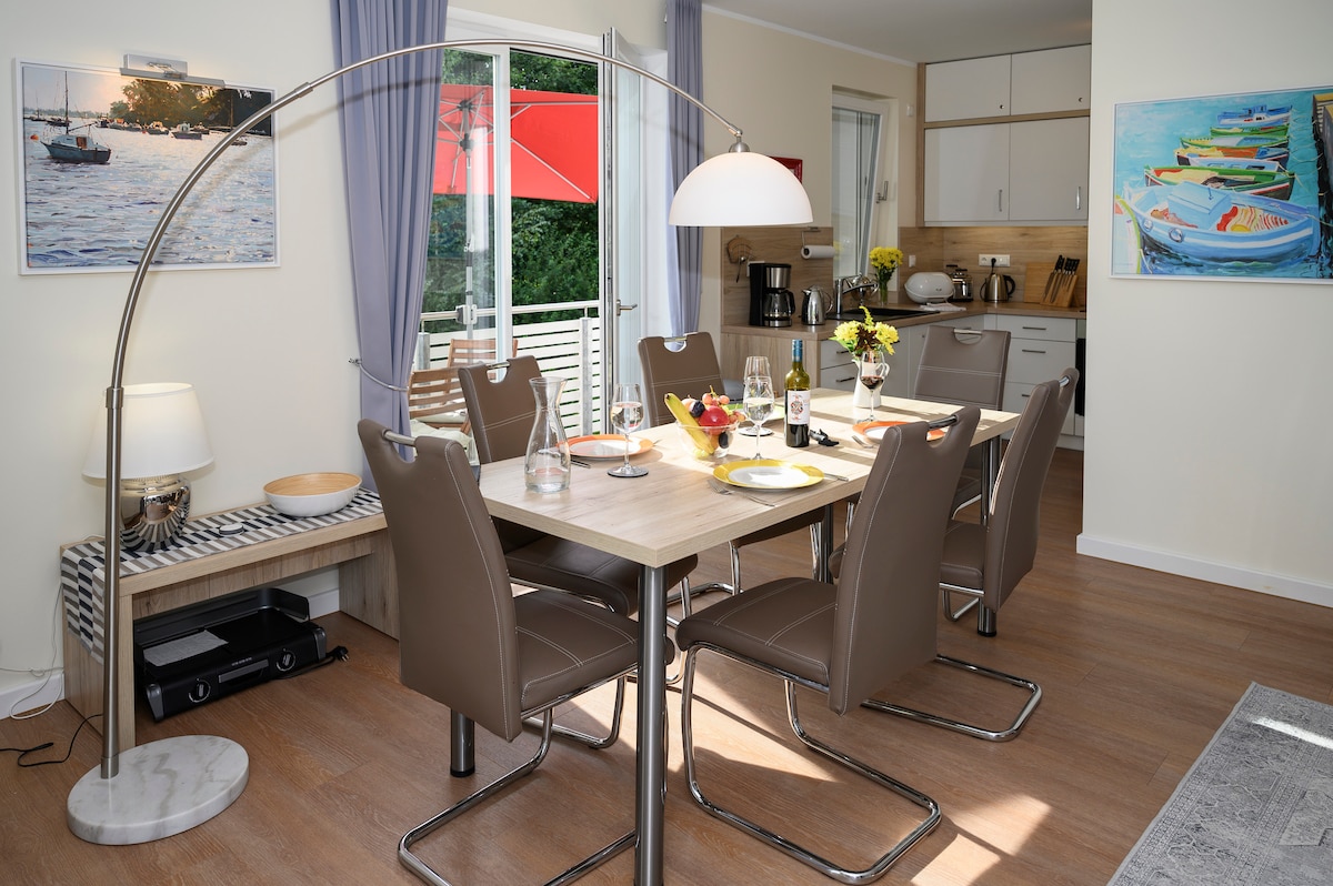 A bright dining area is furnished with a rectangular table and six matching chairs. A floor lamp and a decorative bowl enhance the space. Large sliding doors provide access to a balcony, with scenic views visible through the glass. A kitchenette is seen in the background.