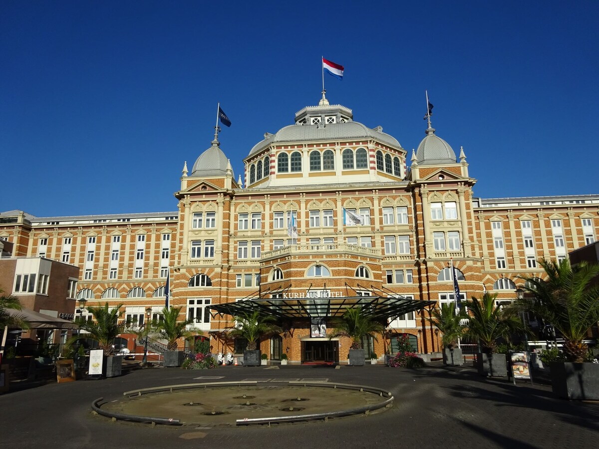 The historic Kurhaus hotel features an impressive façade with distinct architectural details, highlighted by its grand dome and large windows. Flags are visible atop the building, surrounded by palm trees in the foreground, set against a clear blue sky.